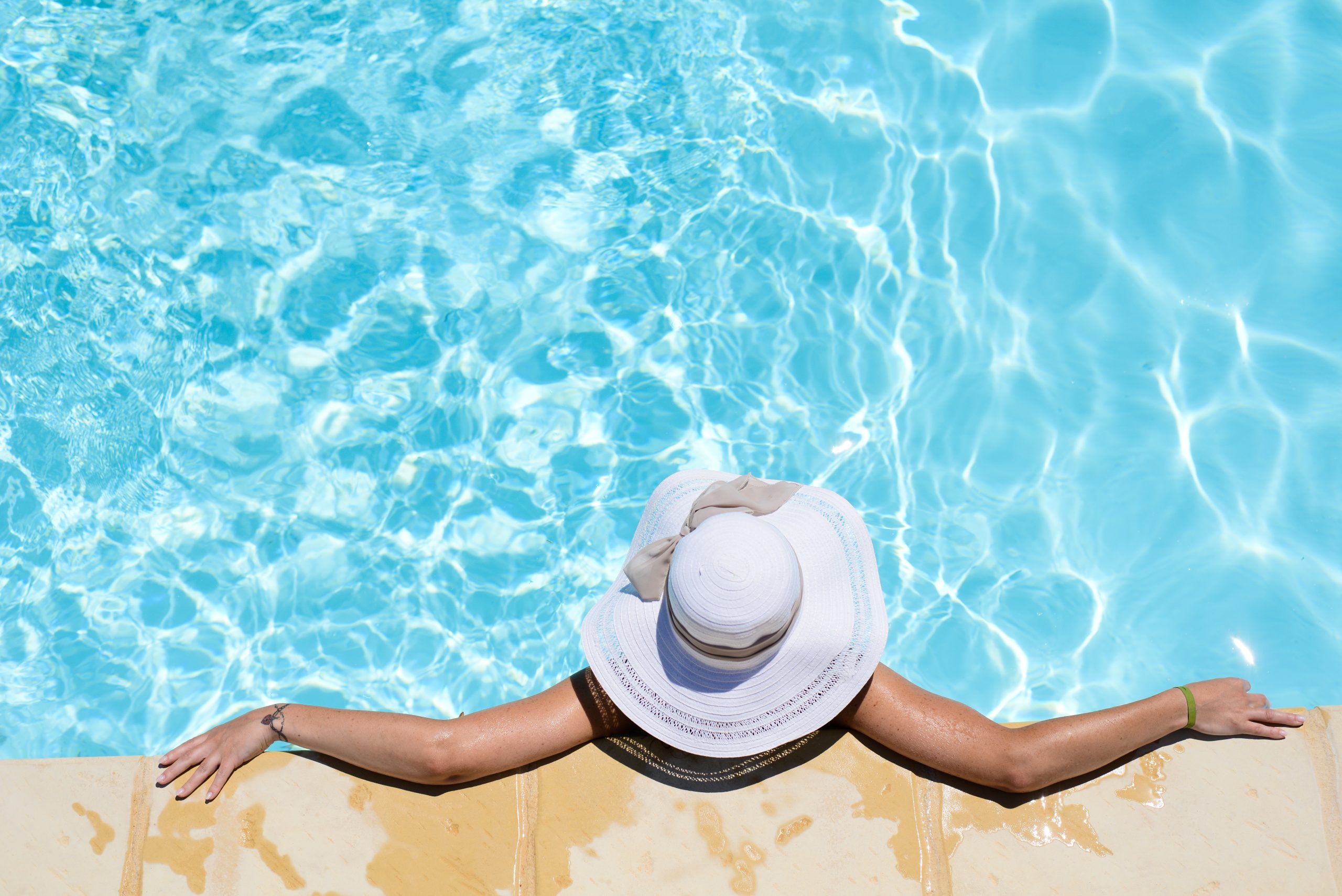 Woman relaxing in backyard pool wearing white hat and arms outstretched